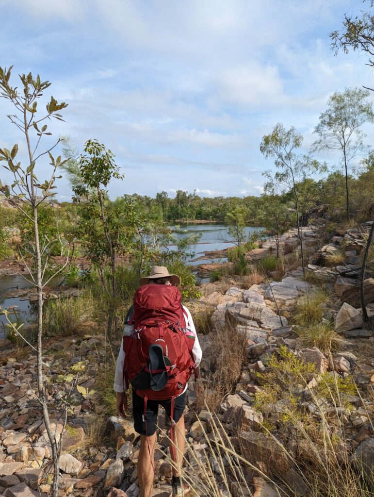 Image with water, rocks, trees, plants and a person hiking the Jatbula Trail in NItmiluk with a red bag and a hat
