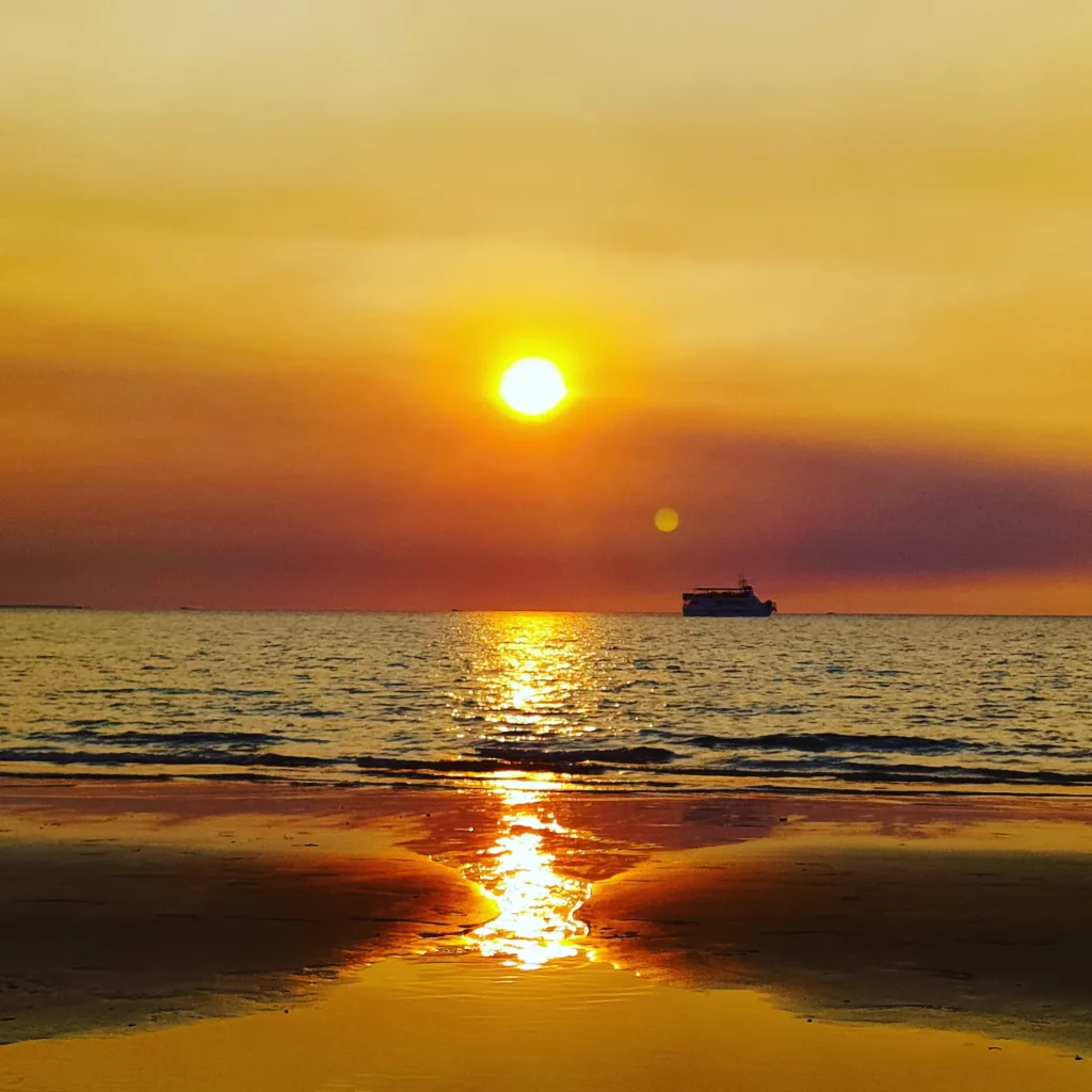 a beautiful sunset at Mindl Beach with orange and yellow skies, a blazing orange sun, dark blue waters, and a dark beach