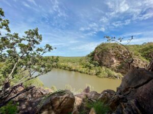 a murky river in between green grass and rock structures with sparse trees surrounding the river and blue skies on the Jatbula Trail