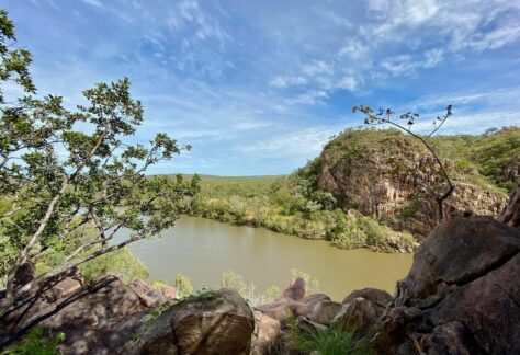 a murky river in between green grass and rock structures with sparse trees surrounding the river and blue skies on the Jatbula Trail