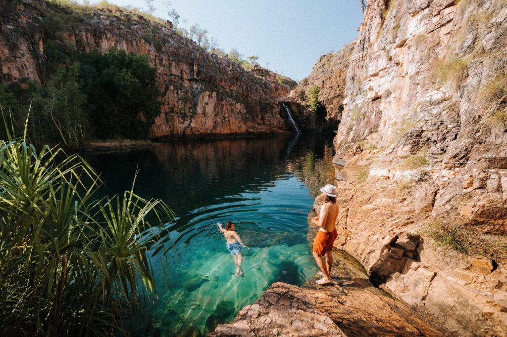 girl swimming to waterfall while man stands on rock